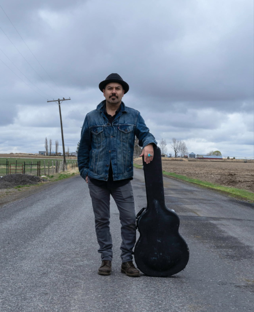 Man with guitar case on rural road.