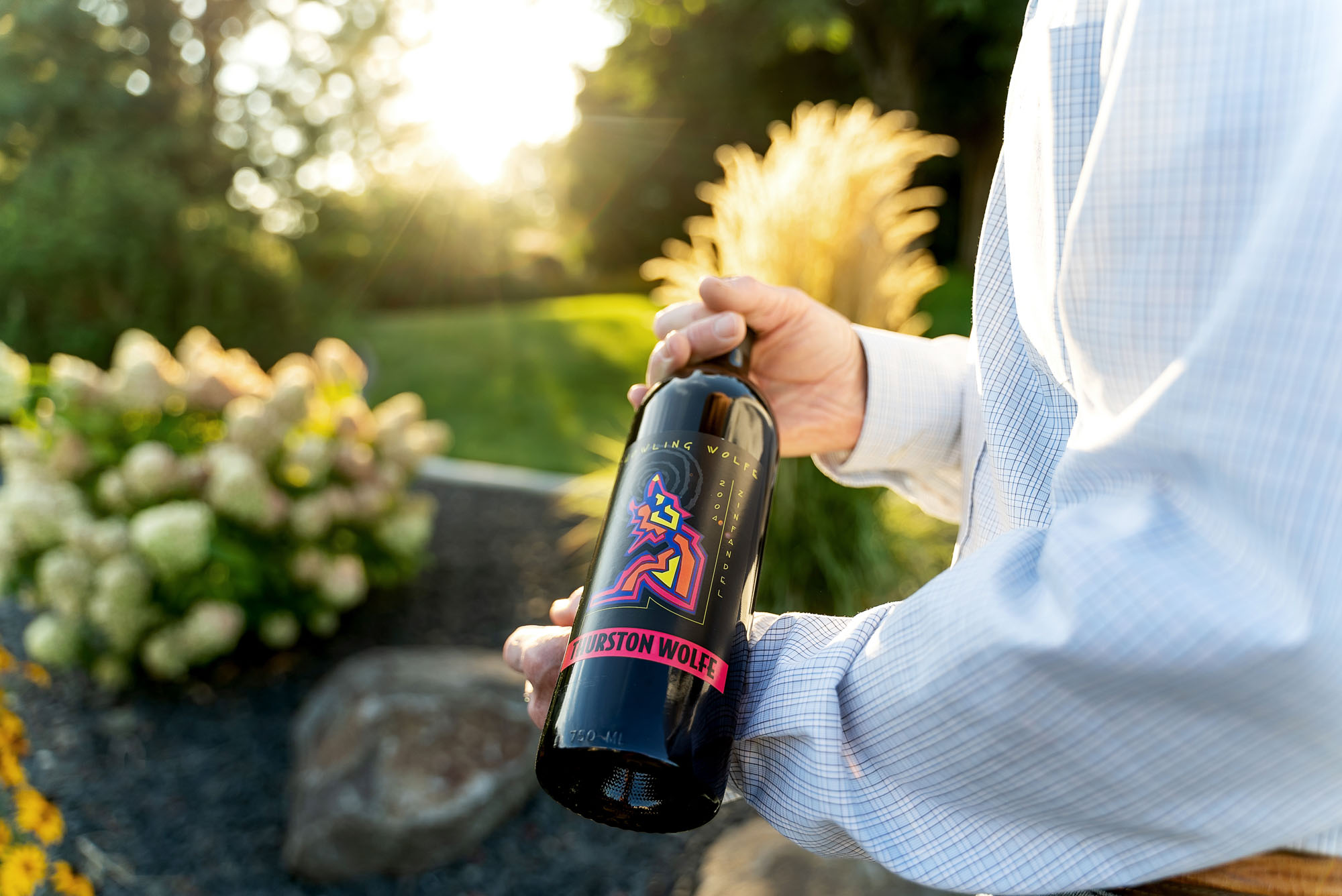 Person holding wine bottle in garden setting.