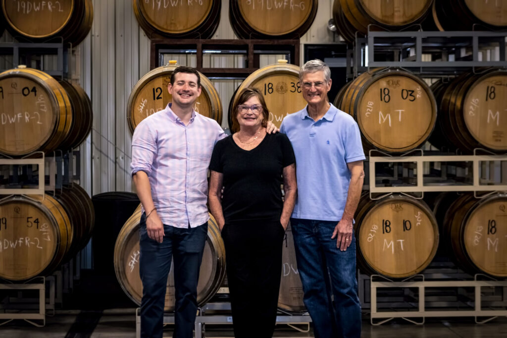 Three people standing in front of wine barrels.